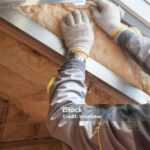 Cropped view on vertical photo with professional worker in protective gloves putting mineral rockwool insulation in wall near windows, behind wooden plank boards. Concept of house under construction